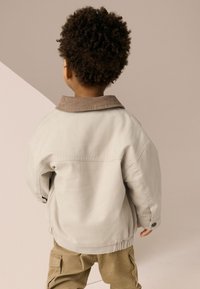 Világos bézs dzseki barna kordbársony gallérral, elasztikus szegéllyel és gombos mandzsettával. Khaki nadrág fölött viselve, hátulnézetből.