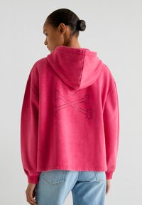 Un sweat à capuche en rose vif avec une texture douce, présentant un design de crâne et os croisés clouté en noir au dos.