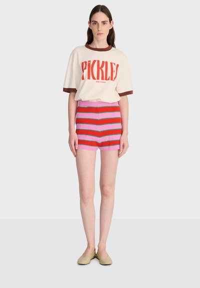 Jeune femme debout, portant un T-shirt beige avec le texte rouge "PICKLES", un short rayé rose et rouge, et des chaussures beiges, fond neutre.