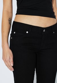 Schwarze Denim-Jeans mit einer figurbetonten Passform, ausgestattet mit silbernen Knopfdetails, vorderen Taschen und einer glatten Textur des Materials.