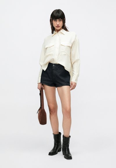 Chemise ample crème clair avec deux poches poitrine, short en denim foncé, bottines noires et petit sac marron. Tissu lisse et coupe décontractée.