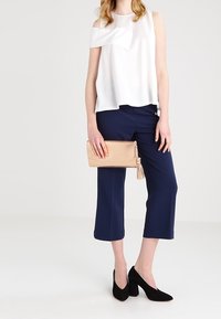 Žena v bílém topu s odhalenými rameny, námořnických zkrácených kalhotách, černých blokových podpatcích, držící béžovou kabelku s třásněmi na jednobarevném pozadí.