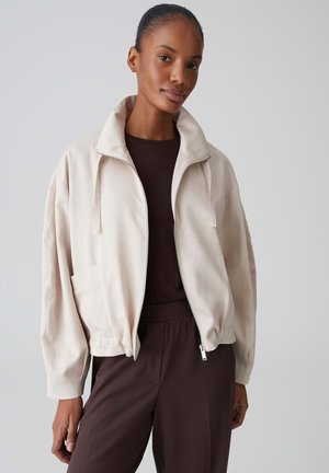 Vrouw met naar achteren gekamd haar, gekleed in een beige jack met ritssluiting over een donkerbruin shirt en bijpassende broek, staand tegen een effen achtergrond.