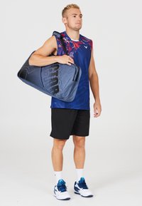 Blå sport duffelväska med svarta detaljer och logotyp, bärs av en person i en blå mönstrad tanktopp och svarta shorts, med vita sneakers.