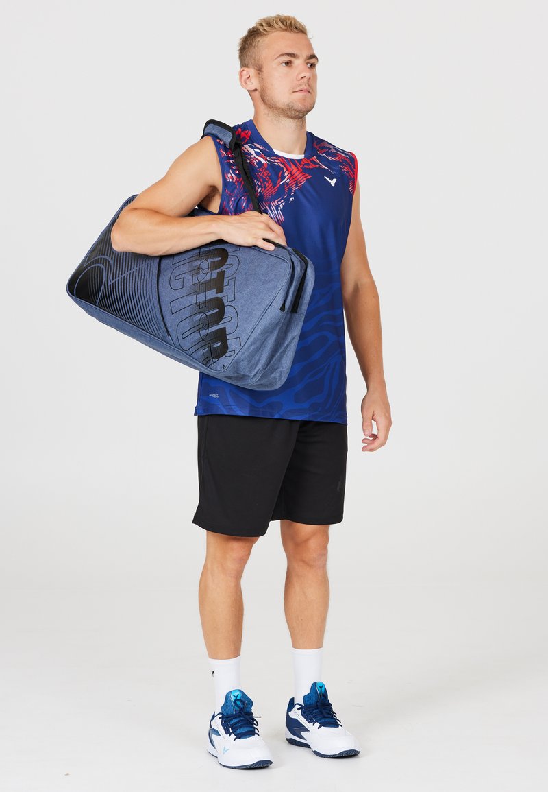 Blå sport duffelväska med svarta detaljer och logotyp, bärs av en person i en blå mönstrad tanktopp och svarta shorts, med vita sneakers.