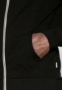 Mano metida en el bolsillo derecho de una sudadera con cremallera negra, con una cremallera blanca visible y una pequeña etiqueta de marca en el bolsillo.