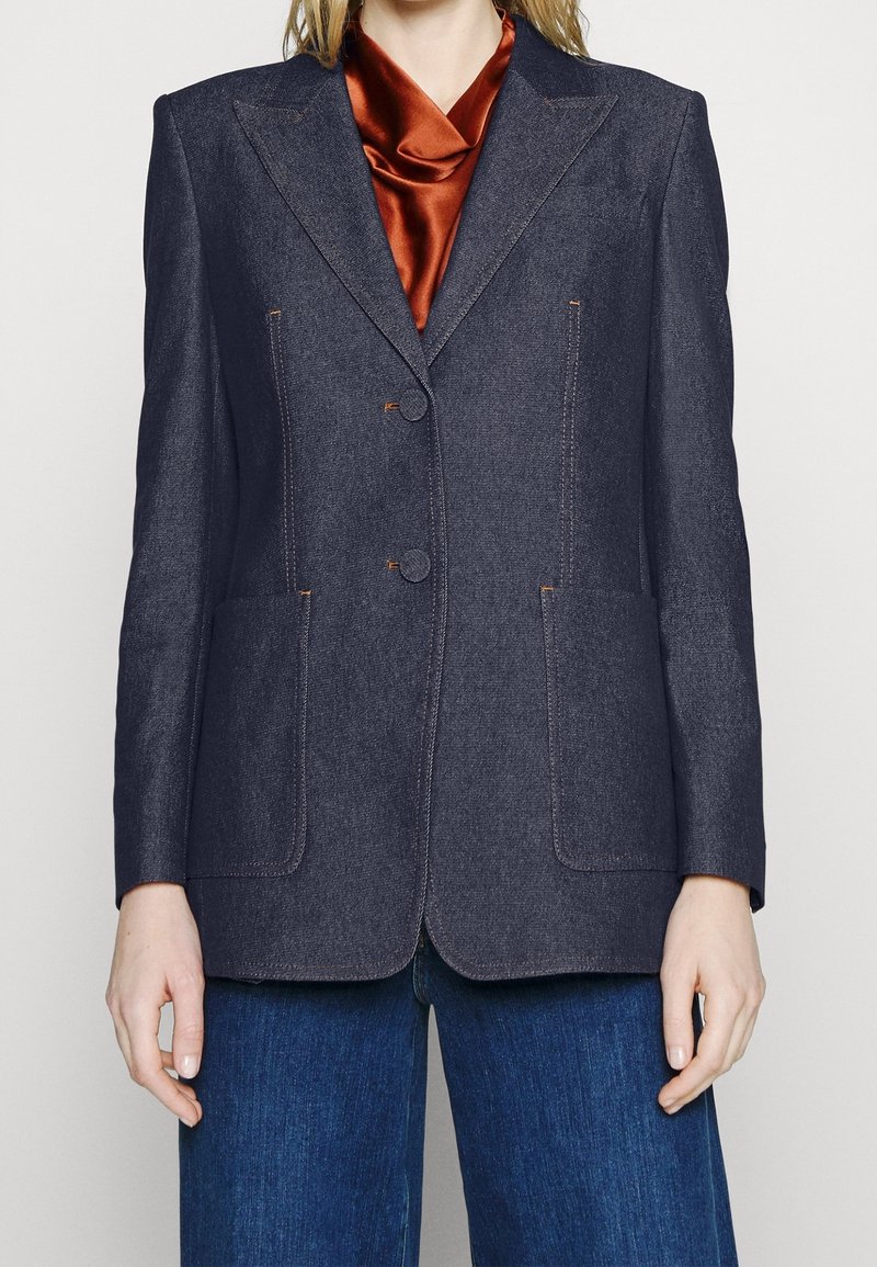 Personne portant un blazer sur mesure gris foncé par-dessus un chemisier en satin couleur rouille et un jean en denim bleu, debout devant un fond uni.