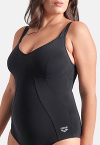 Sort badedragt i ét stykke lavet af glat stof med tynde stropper, V-udskæring og diskrete syningsdetaljer. Indeholder "arena"-logoet.