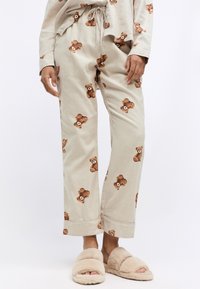 Beige pyjamabroek met een bruin berenprint, gemaakt van zachte stof, met een elastische tailleband met trekkoord en opgerolde boorden. Gepaard met fluffy slippers.