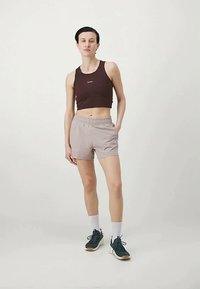 Moteris stovi su viena ranka įsikišusi į bežinius šortus, vilkinti tamsiai rudą trumpą palaidinę, baltas kojines ir tamsiai žalias sportines batus.