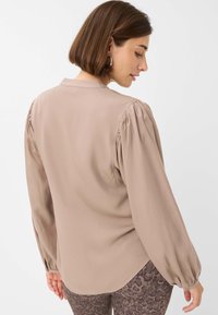 Blusa taupe con maniche lunghe a sbuffo, tessuto morbido, colletto con bottoni sul retro e orlo curvo. Indossata con leggins marroni a fantasia.