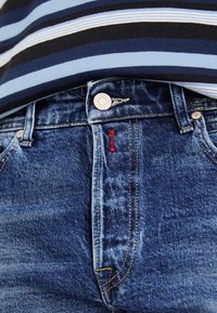 Jeans de mezclilla en lavado medio con un diseño de cinco bolsillos, botón plateado y detalle de etiqueta roja en el lateral. Llevados con una camisa a rayas.