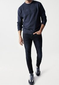 Uomo con una felpa blu scuro con il testo "SALSA JEANS", jeans neri aderenti e sneakers bianche e blu navy in piedi su uno sfondo bianco.