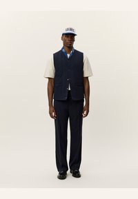 Navy quilted vest, beige short-sleeve shirt, navy trousers, and black shoes. Cap with blue and white text. Notable layering and colors.