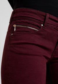 Burgunderfärgade jeans med en följsam textur, som har en liten ficka med dragkedja, synlig sömnad och en metallisk dragkedja. Mellanhög midja.