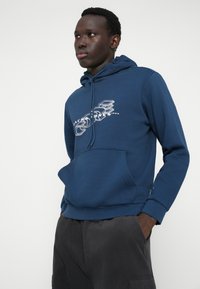 Tumši zila kapučjaka jakas, kas izgatavota no mīksta auduma, ar priekšējo kabatu, ar auklu regulējamu kapuci un grafisku apdruku uz krūtīm. Apvienota ar tumšiem biksēm.