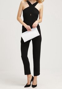 Black jumpsuit with a crisscross design and keyhole detail, paired with a white clutch and black pointed-toe heels. Smooth texture, fitted shape.
