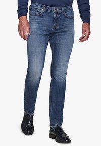 Jeans in denim slim-fit di colore blu scuro con finitura sfumata, dotati di cinque tasche e chiusura con bottone. Indossati con scarpe nere lucide.