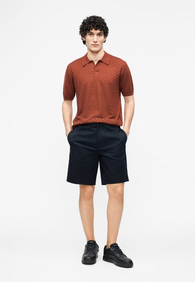 Νέος άνδρας όρθιος με τα χέρια στις τσέπες, φοράει κοντομάνικο πόλο σε σκουρό κόκκινο χρώμα, μαύρο σορτς και μαύρα αθλητικά παπούτσια μπροστά σε απλό φωτεινό φόντο.