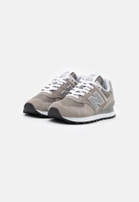 Pár šedých a bílých tenisek New Balance 574 s bílými tkaničkami, síťovanými panely, semišovými překryvy a černými gumovými podrážkami na bílém pozadí.