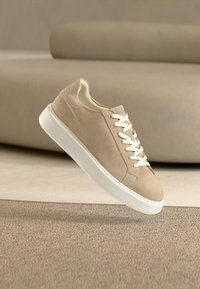 Μπεζ σουέντ αθλητικό παπούτσι με λευκά κορδόνια και χοντρή λευκή σόλα που αιωρείται πάνω από μια υφασμάτινη μπεζ μοκέτα σε ουδέτερο εσωτερικό χώρο.