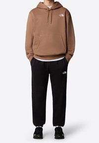 Brun huvtröja med kenguruficka och vit logotyp, i kombination med svarta träningsoverallsbyxor med logotyp, och svarta sneakers.