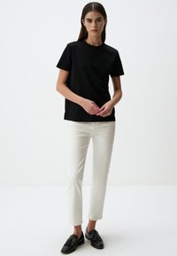 Zwart T-shirt met korte mouwen en ronde hals, gecombineerd met witte fitted broek en zwarte instappers, gedragen tegen een effen achtergrond.
