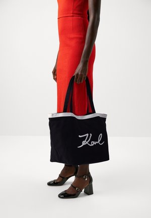 Persona con un vestido rojo sosteniendo un gran bolso negro con ribete blanco y con "Karl" escrito en blanco, vistiendo zapatos de tacón alto negros.