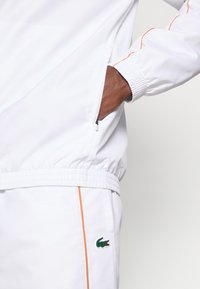 Λευκή αθλητική ζακέτα με πορτοκαλί ρίγες, ελαστικούς καρπούς και πλευρική τσέπη. Διαθέτει πράσινο λογότυπο Lacoste στην αριστερή πλευρά.