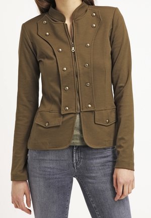 Veste marron style militaire avec boutons métalliques double rangée et poches à rabat portée sur un top vert et un jean bleu.