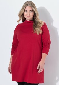 Donna con lunghi capelli biondi che indossa grandi orecchini a cerchio rossi e un vestito tunica rosso con maniche lunghe, in piedi contro uno sfondo bianco.