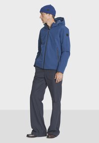 Blue zip-up jacket with a hood, featuring two zippered pockets and a textured finish, paired with dark pants and a blue knitted beanie.