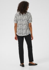 Blouse à manches courtes avec un motif abstrait noir et blanc, associée à un pantalon noir et des chaussures plates noires. Le tissu semble lisse et léger.
