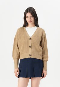 Beige strikket cardigan med v-hals, lange ermer og tre knapper foran. Har ribbestrikket tekstur og kort design.