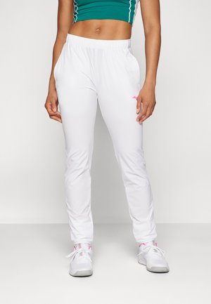 Sportliche Person trägt weiße Trainingshose, weiße Sneakers mit pinkfarbenen Akzenten und ein grünes kurzes Oberteil mit weißen Paspeln, steht vor weißem Hintergrund.