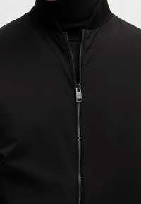 Musta bomber-tyylinen takki, joka on valmistettu sileästä materiaalista. Takissa on pystykaulus ja etuvetoketjukiinnitys, jossa on hopeinen metallinen vetonuppi.