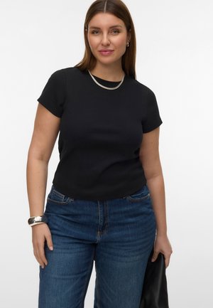 Frau mit glatten braunen Haaren trägt ein schwarzes kurzärmeliges Oberteil, blaue Jeans, eine silberne Halskette und ein Armband und hält eine schwarze Ledertasche.