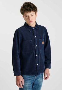 Navy corduroy shirt met knoopsluiting, twee voorkruiszakken en een klein logo-opdruk. Draagt bij blauwe denim jeans.