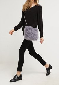 Szara torebka crossbody z sztucznego futra z silwerską łańcuszkową rączką, prostokątnego kształtu, o miękkiej teksturze, noszona przez osobę w czarnym ubraniu.