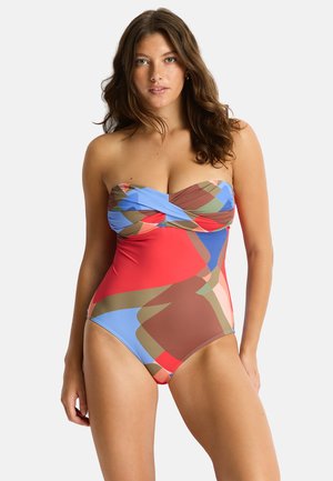 Femme portant un maillot de bain une pièce sans bretelles avec des motifs géométriques abstraits en rouge, bleu, marron et vert, debout devant un fond uni.