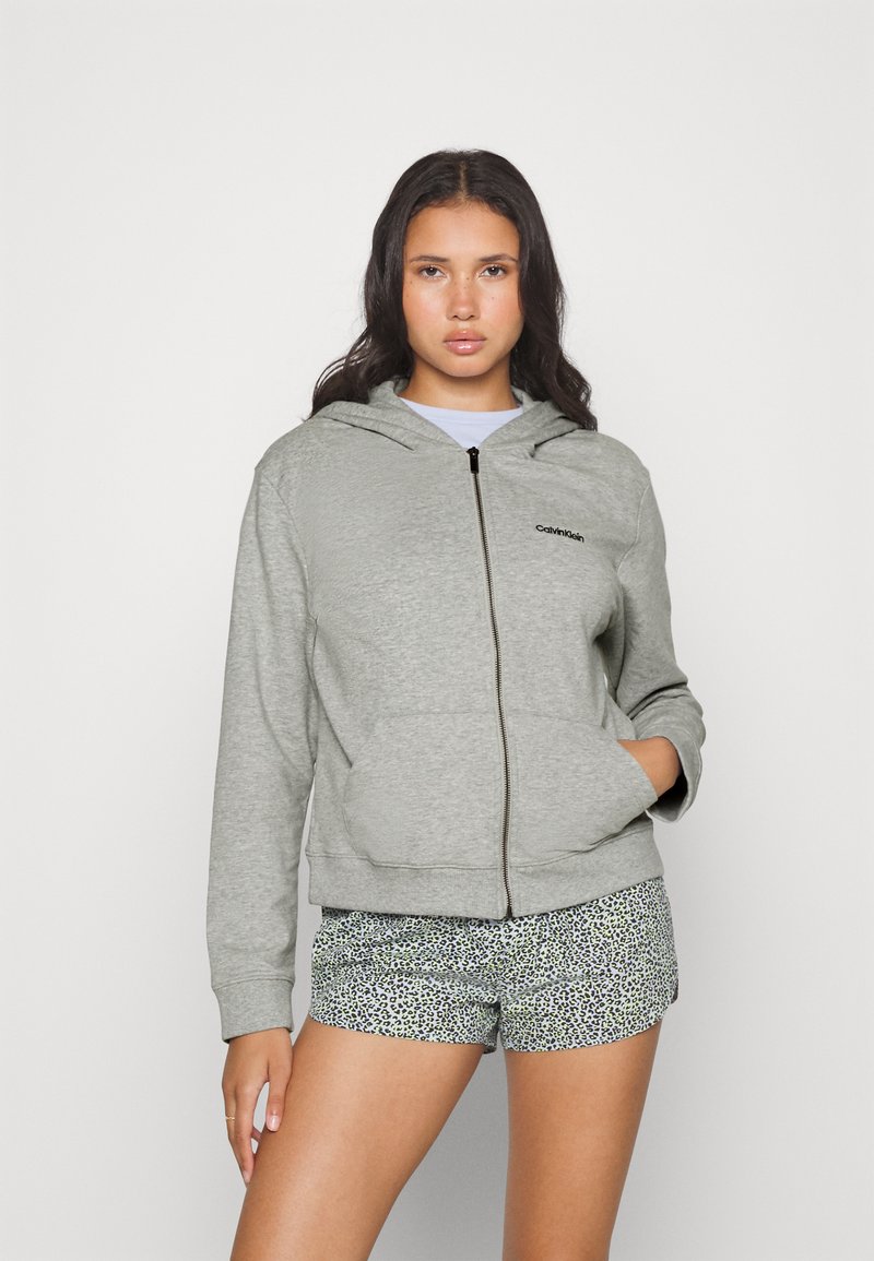 Sudadera con cremallera gris de textura suave, con bolsillo canguro y capucha; presenta un logo negro en el lado izquierdo del pecho. Combinada con pantalones cortos estampados.