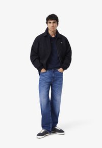 Marinblå zip-up jacka över en marinblå polo, kombinerad med tvättade blå jeans och marinblå sneakers med vita snörningar.