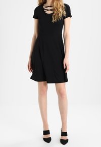 Robe noire à manches courtes et décolleté en croix. Jupe évasée et ourlet jusqu'aux genoux. Portée avec des chaussures à talons hauts noires.