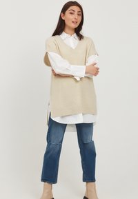 Gilet en tricot beige avec un col en V, superposé à une chemise blanche à boutons. Porté avec un jean bleu et des bottines beige.