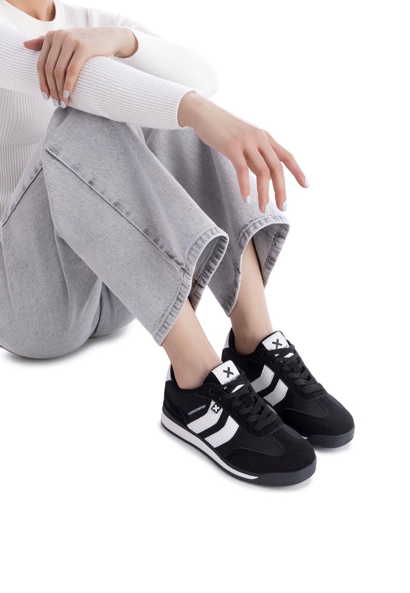Sneakers in bianco e nero con un upper in suede, dotati di chiusura con lace, strisce bianche e suola spessa in gomma, abbinati a pantaloni grigi a tre quarti.