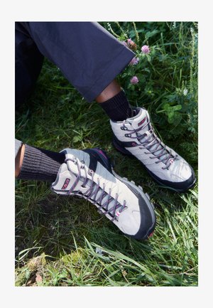 Vaaleanharmaat vaelluskengät mustilla yksityiskohdilla, vaaleanpunaiset nauhat ja teksturoidut pohjat, aseteltuna ruohoon tummien ribattujen sukkien kanssa.