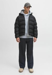 Mężczyzna w czarnej kurtce puchowej noszący szary hoodie, czarne spodnie, beżowe buty oraz szary czapkę stoi na tle gładkiej białej ściany.