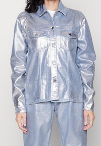 Giacca di jeans metallica blu chiaro con maniche lunghe, due tasche sul petto, bottoni argentati e un design colletto classico. Texture liscia e lucida.