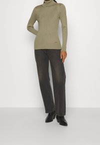 Oliwkowy sweter z prążkowanym kołnierzem i długimi rękawami, zestawiony z ciemnoszarymi spodniami typu wide-leg oraz czarnymi botkami na obcasie.