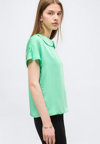 Jeune femme portant un chemisier vert clair à manches courtes avec un col arrondi et un pantalon noir, debout de profil devant un fond uni.